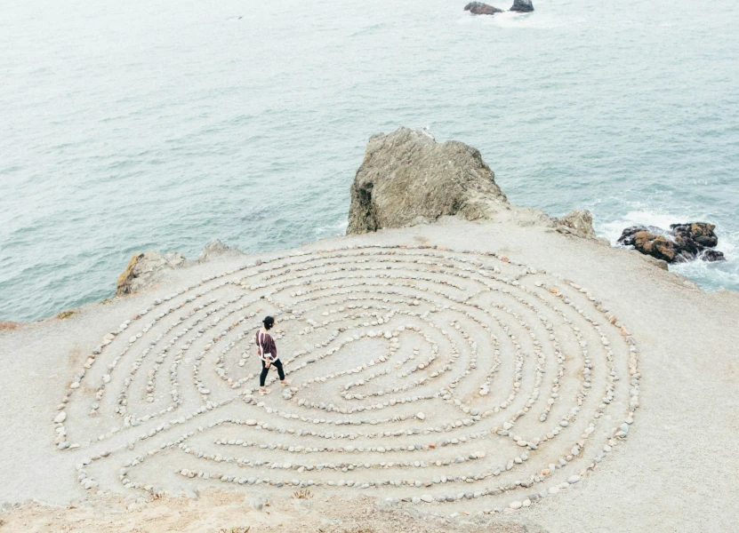 stein-labyrinth-psychotherapeutische-beratung Zu sehen ist ein Stein-Labyrinth. Eine Frau geht durch das Labyrinth. Das Labyrinth ist vor einem Felsvorsprung mit angrenzendem Meer. Die psychotherapeutische Beratung hilft durch das eigene Labyrinth zu finden.